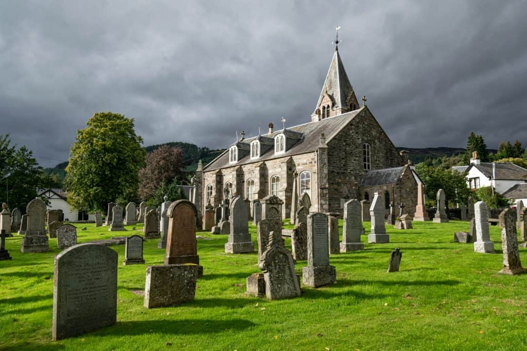 A historic stone church with a tall spire, surrounded by a graveyard with various tombstones on a grassy area under a cloudy sky.