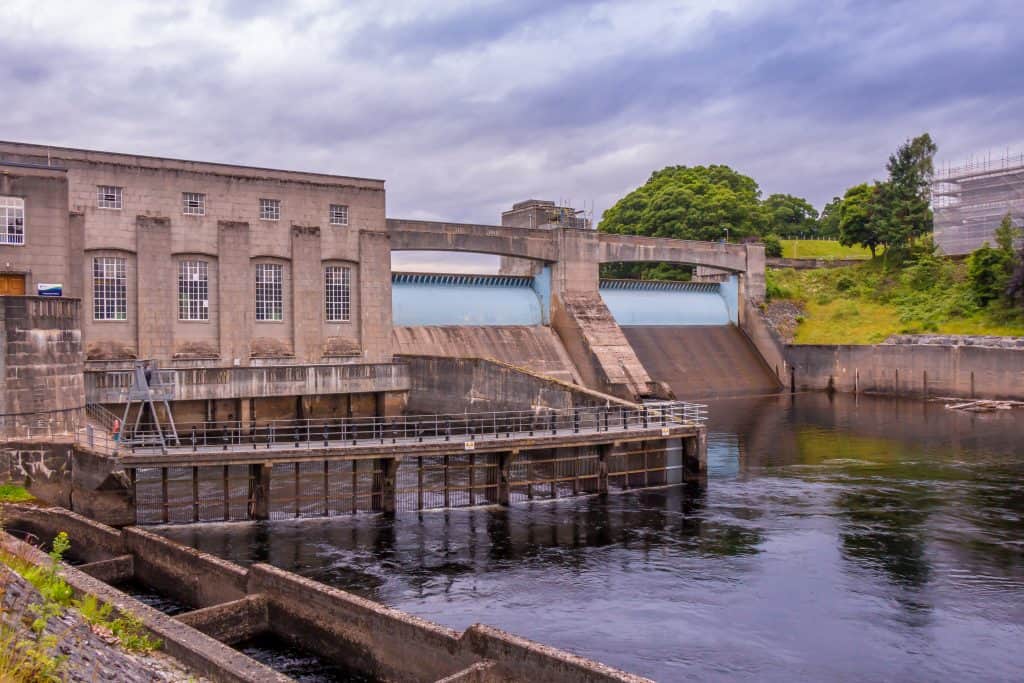 A hydroelectric power station at Pitlochry beside a river with a dam structure, surrounded by greenery and a cloudy sky.