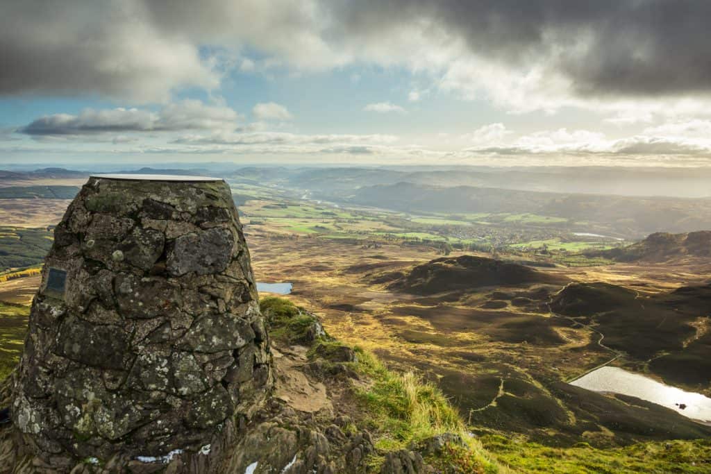 Stone trig point on a hilltop with a wide view of a green valley, distant hills, patches of sunlight, and scattered clouds in the sky.