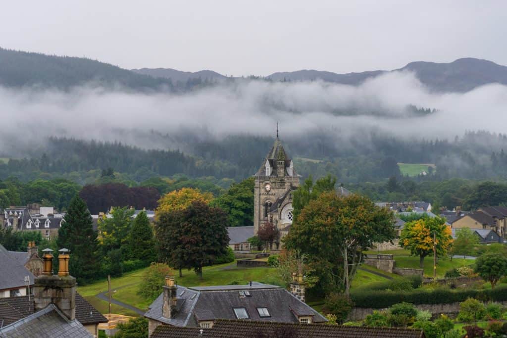 Scenic landscape in Pitlochry with a clock tower, trees, and rooftops, set against a backdrop of misty hills and forests.