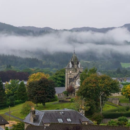 Scenic landscape in Pitlochry with a clock tower, trees, and rooftops, set against a backdrop of misty hills and forests.