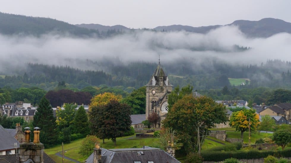 Scenic landscape in Pitlochry with a clock tower, trees, and rooftops, set against a backdrop of misty hills and forests.