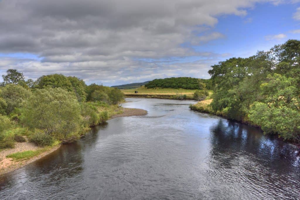 A calm river flows through a lush green landscape under a partly cloudy sky near Aviemore.
