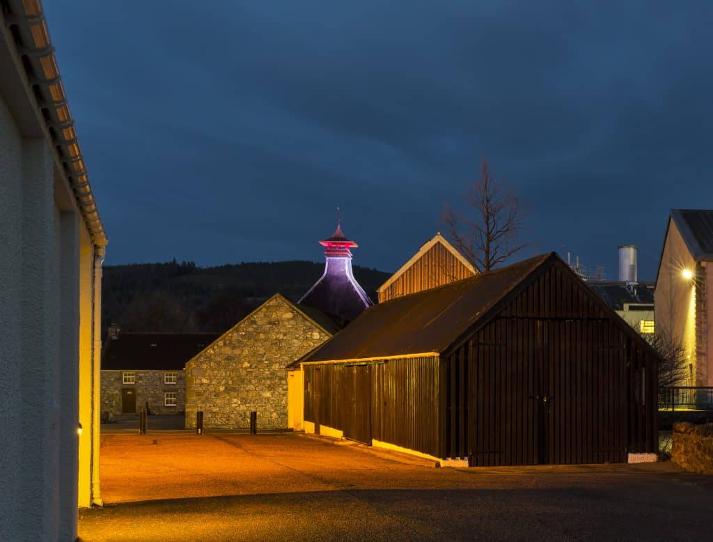 Evening view of a distillery courtyard with stone and wooden buildings, and a tower illuminated by purple and red lights against a dark sky.