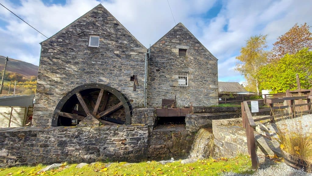 Historic stone watermill with large wooden wheel, surrounded by autumn trees and having a small stream flowing beside it on a sunny day.