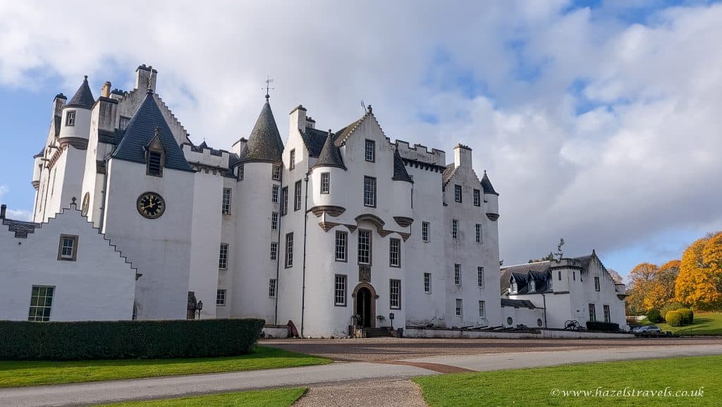 Blair Castle, a white historic building with multiple turrets and chimneys, under a partly cloudy sky. Surrounded by greenery and an autumn-colored tree on the right.