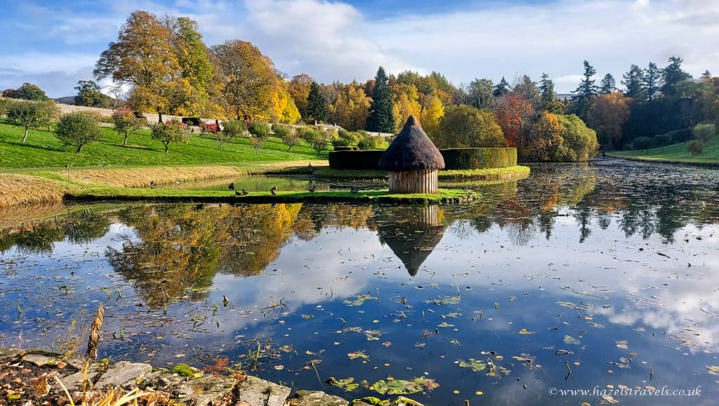A small thatched hut on a reflective lake surrounded by autumn trees and greenery, under a partly cloudy sky.