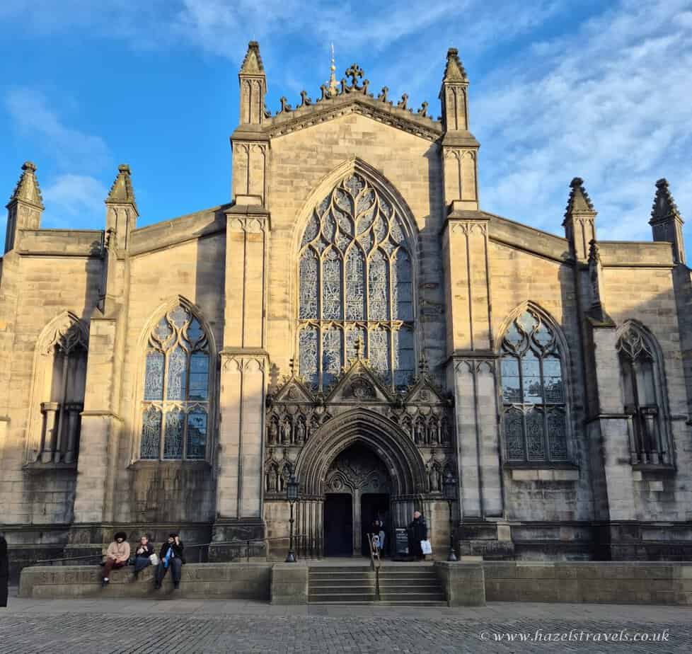 The facade of a large Gothic-style cathedral in Edinburgh with pointed arches and elaborate stained glass windows under a blue sky. Several people are seated or standing outside.