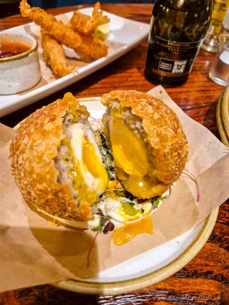 A halved Scotch egg on a plate, showing a soft, runny yolk. In the background, a dish with fried shrimp and a bottle of Prosecco are visible.