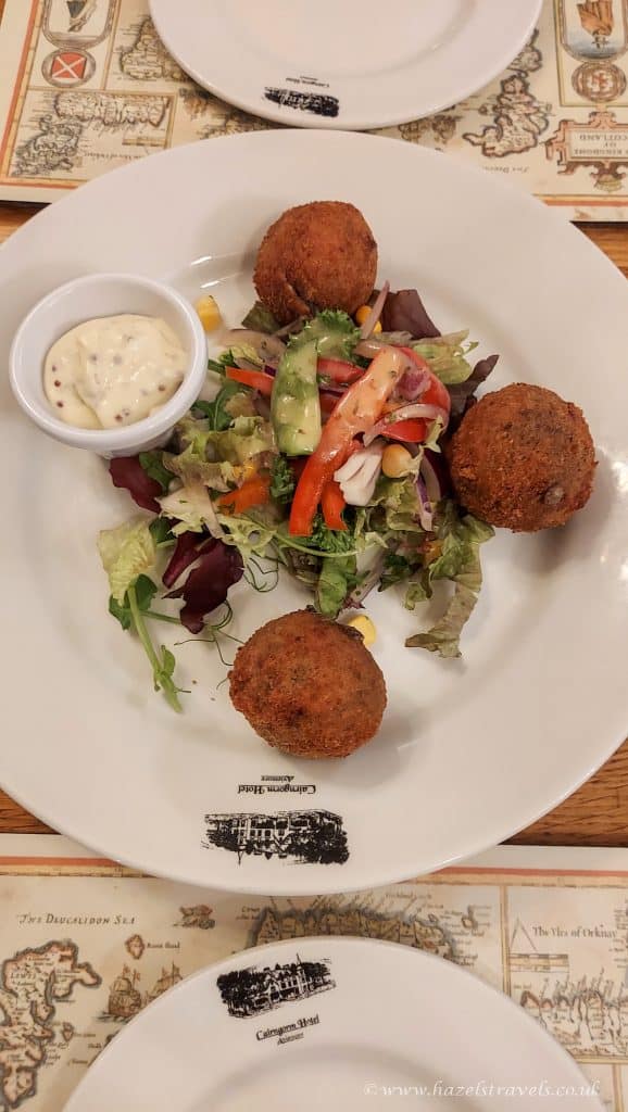 A plate of haggis bonbons (a food of scotland) mixed salad with cucumber, tomato, and onion, served with three breaded croquettes and a small dish of tartar sauce.