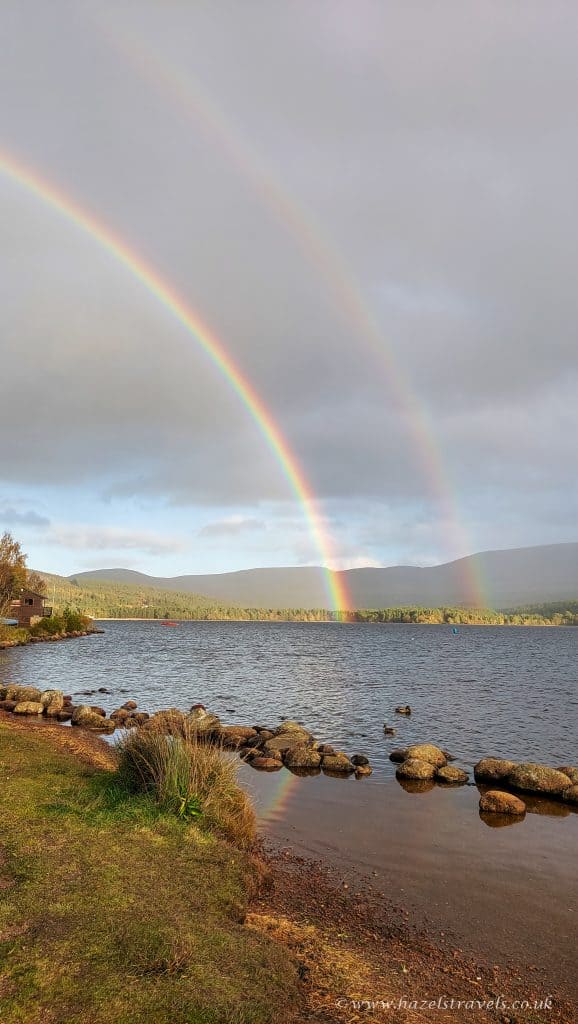 A double rainbow arcs over a calm lake with a rocky shore, under a cloudy sky.