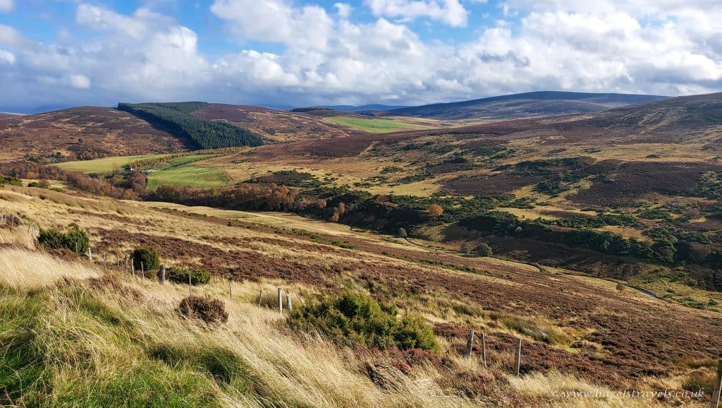 A panoramic view of a hilly landscape with patches of grass, shrubs, and distant trees under a partly cloudy sky.