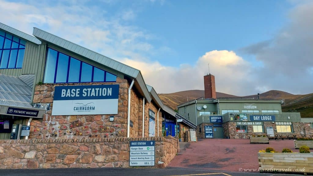 Exterior of Cairngorm Mountain Base Station, featuring a stone building with signs for various facilities, under a partly cloudy sky.