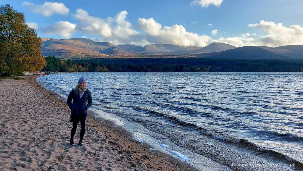 A person walks along a sandy lakeshore with mountains and a forest in the background under a partly cloudy sky.