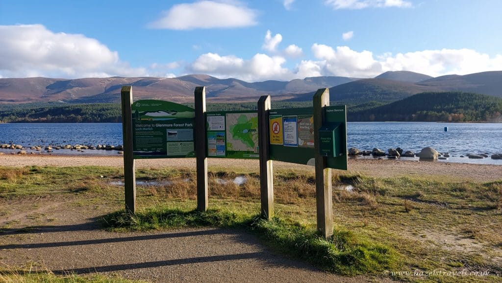 Information boards at the edge of a lake with distant mountains under a partly cloudy sky.