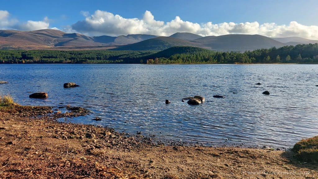 Loch Morlich with visible rocks in the water, surrounded by a forest with distant mountains under a blue, cloudy sky.