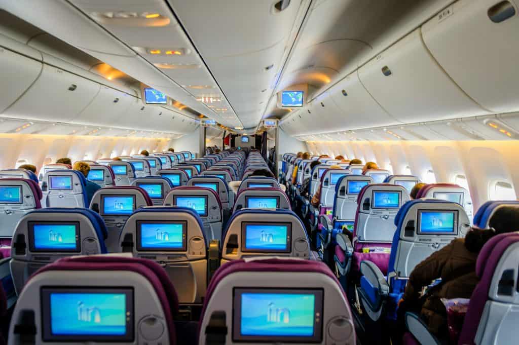 Interior of a commercial airplane cabin, showing rows of passenger seats equipped with personal screens and overhead luggage compartments.