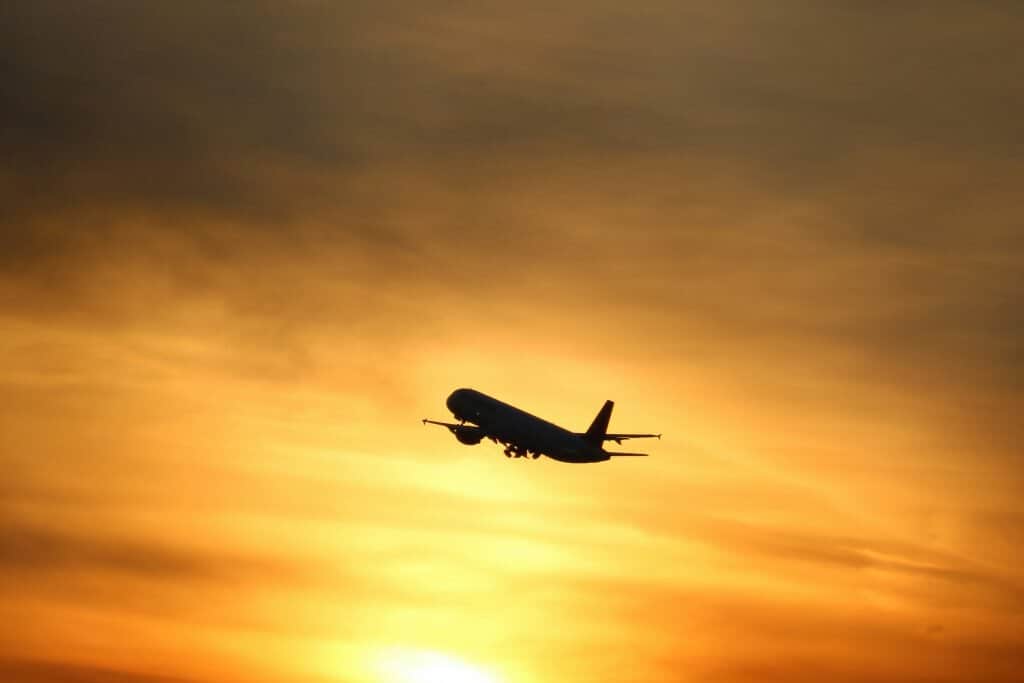 Airplane silhouette against an orange sunset sky, climbing upward.