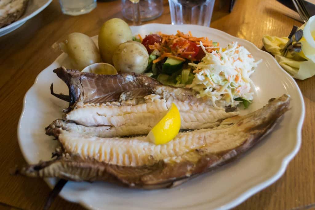 A plate with sliced smoked fish, a lemon wedge, boiled potatoes, and a fresh salad including tomatoes, cucumbers, and coleslaw.