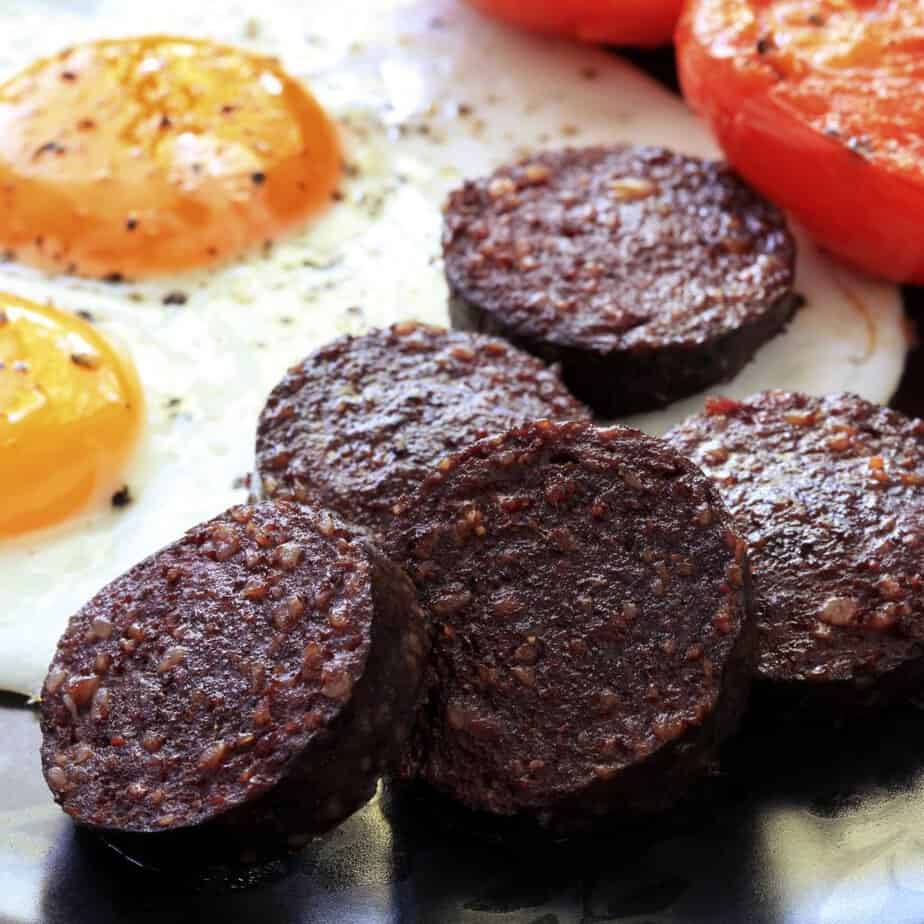 Breakfast plate with two sunny-side-up eggs, slices of black pudding, and roasted tomatoes.