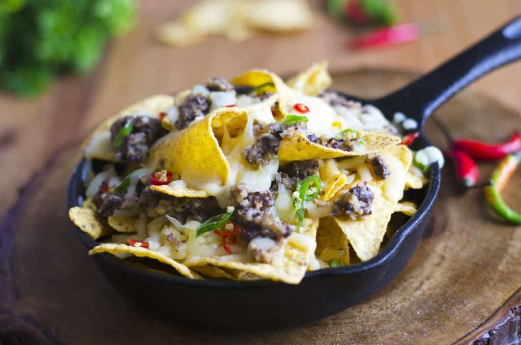 A skillet filled with nachos topped with melted cheese, ground meat, and sliced red chilies, placed on a wooden surface.