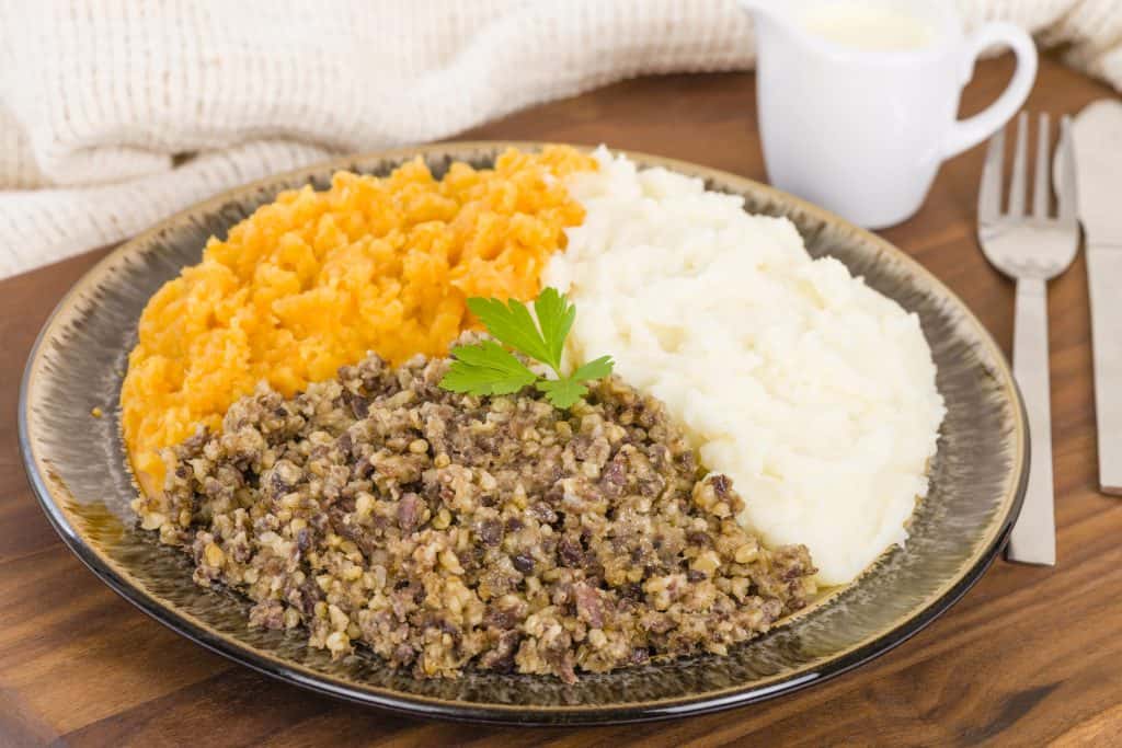 A plate with three sections: mashed sweet potatoes, mashed potatoes, and a portion of haggis, garnished with a parsley sprig.