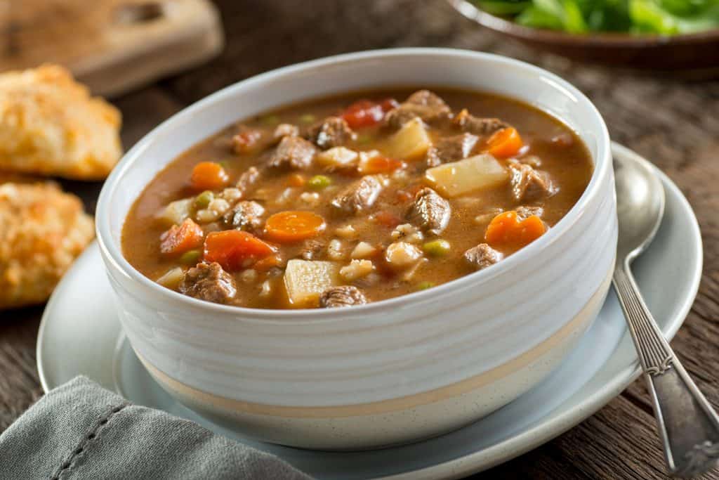A bowl of beef stew with carrots, potatoes, and peas, served with a spoon on the side on a wooden table.