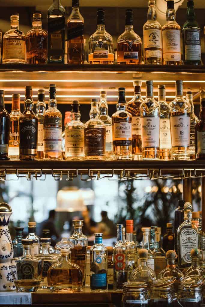 A variety of liquor bottles, including whiskey and tequila, are displayed on illuminated shelves in a bar setting.