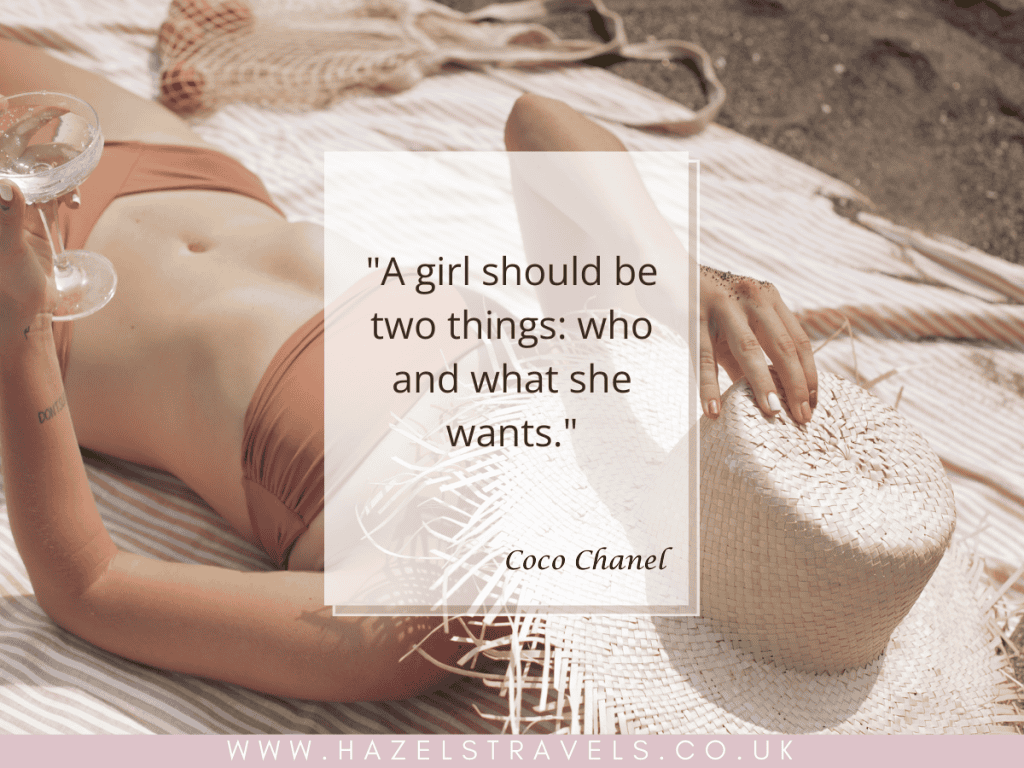 Woman in a bikini lying on a towel at the beach, holding a hat and glass. Coco Chanel quote about solo female travel overlays image.