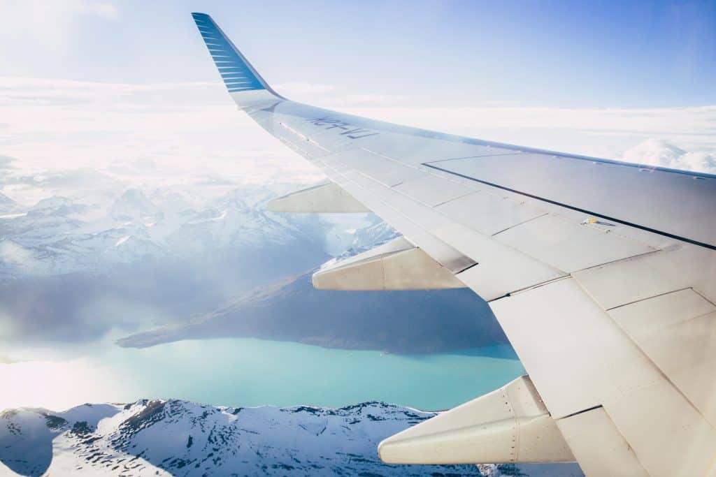 View from airplane window showing a wing over snowy mountains and a turquoise lake under a clear sky.