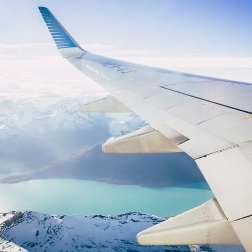 View from airplane window showing a wing over snowy mountains and a turquoise lake under a clear sky.