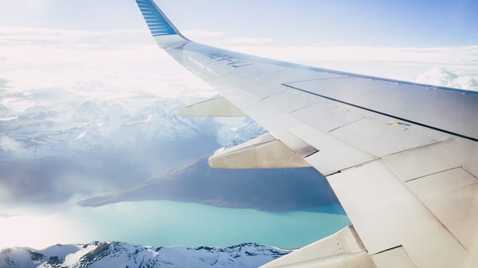 View from airplane window showing a wing over snowy mountains and a turquoise lake under a clear sky.