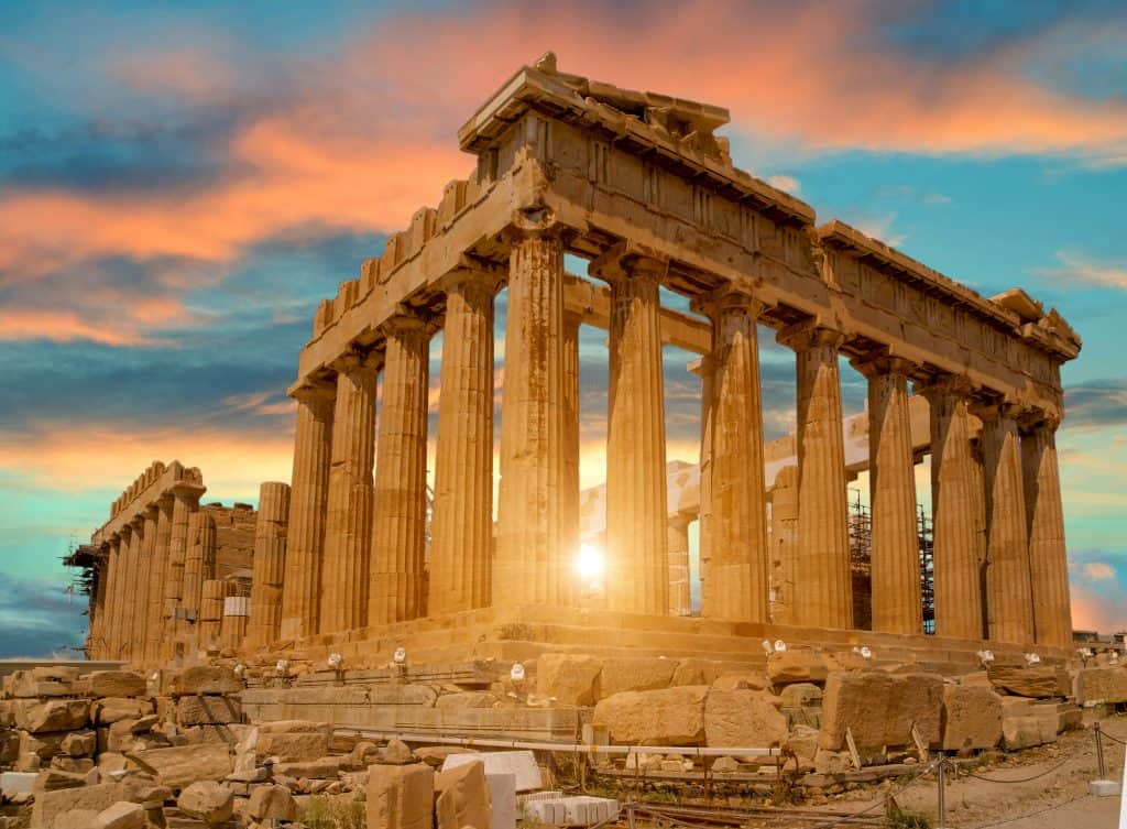 The Parthenon at sunset, with sun rays peeking through its ancient columns, under a partly cloudy sky.