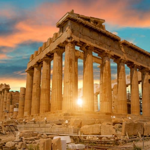 The Parthenon at sunset, with sun rays peeking through its ancient columns, under a partly cloudy sky.