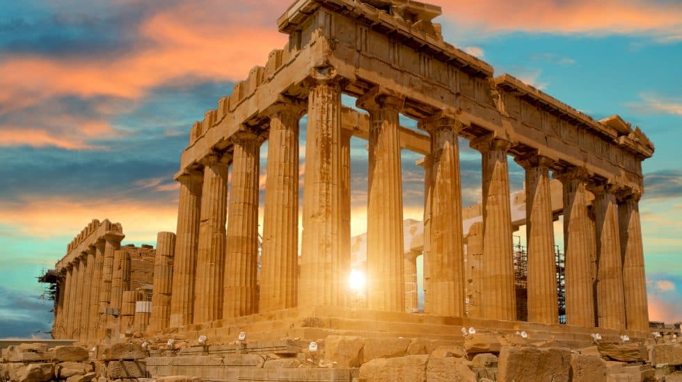 The Parthenon at sunset, with sun rays peeking through its ancient columns, under a partly cloudy sky.