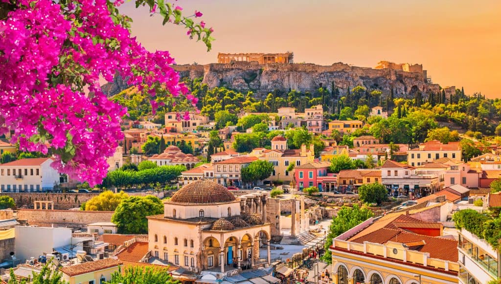 View of Athens with vibrant pink flowers in the foreground, city buildings, and the ancient Acropolis atop a hill under a warm, colorful sky.