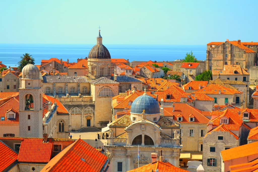A scenic view of Dubrovnik's old town with its iconic red-tiled roofs, historic domes, and bell towers under a clear blue sky, overlooking the Adriatic Sea.