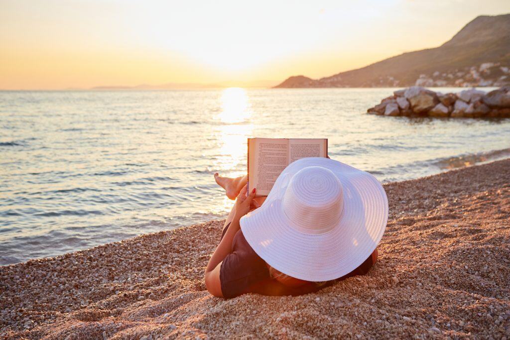 Person reading a book on a pebble beach at sunset, wearing a large white sun hat. The calm sea and distant hills are visible in the background.