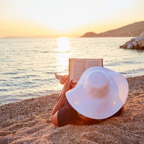 Person reading a book on a pebble beach at sunset, wearing a large white sun hat. The calm sea and distant hills are visible in the background.