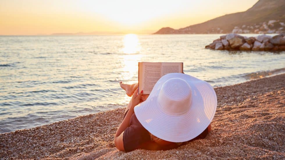 Person reading a book on a pebble beach at sunset, wearing a large white sun hat. The calm sea and distant hills are visible in the background.