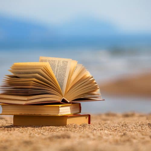An open book on top of two closed books sits on sandy beach with a blurred ocean background.