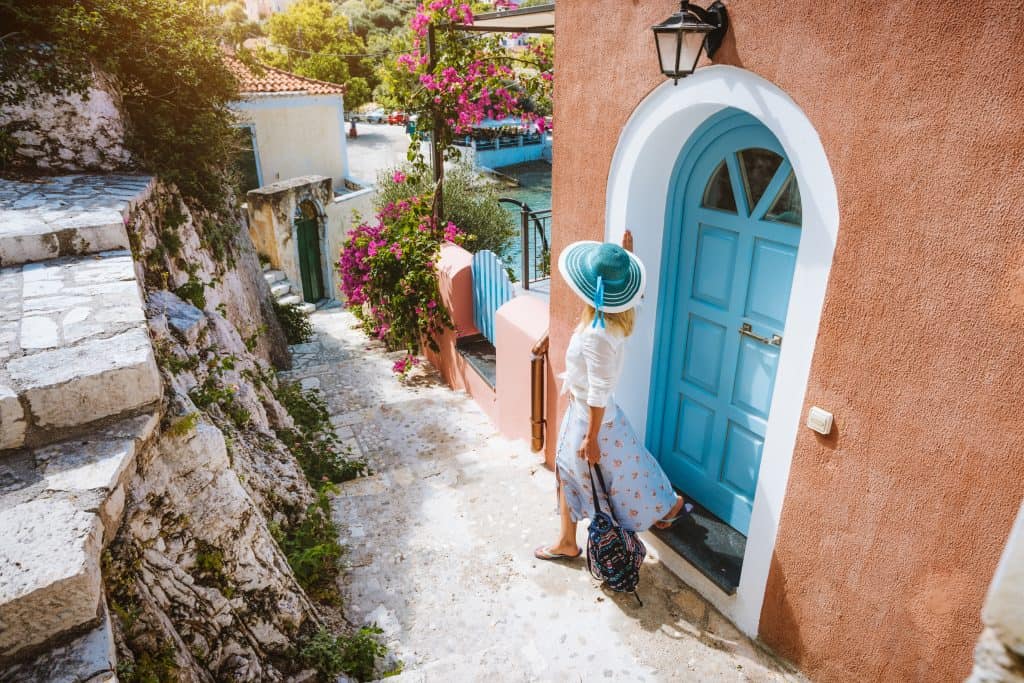 A woman in a sunhat stands by a blue door on a narrow, scenic street lined with colorful buildings and vibrant flowers.