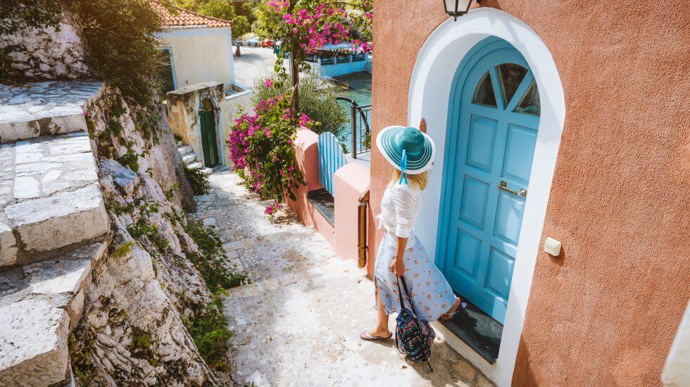 A woman in a sunhat stands by a blue door on a narrow, scenic street lined with colorful buildings and vibrant flowers.