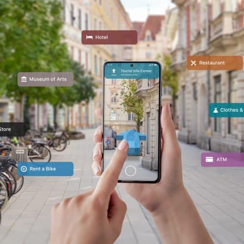 Hands hold a smartphone showing augmented reality navigation in a city street. Icons display nearby locations: hotel, restaurant, museum, mobile store, bike rental, fashion, ATM.