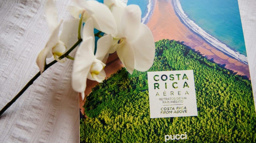 A book titled "Costa Rica Aérea" with an aerial view of a coastline on the cover, placed beside a white orchid on a textured white surface.