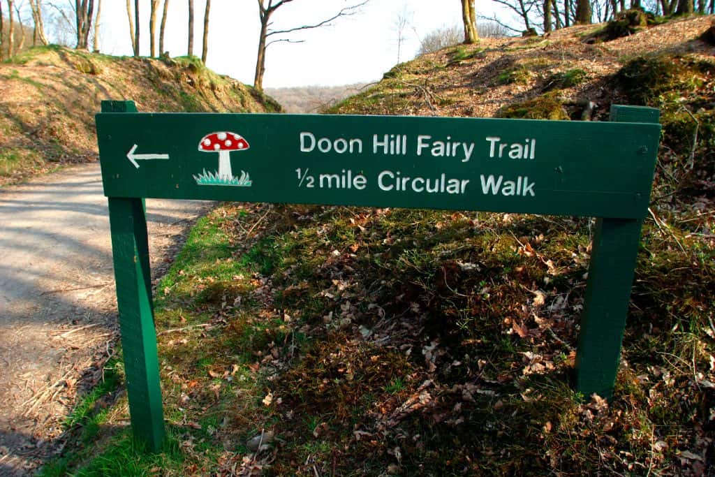 Green sign in wooded area reads "Doon Hill Fairy Trail, ½ mile Circular Walk" with an arrow pointing left and a red mushroom illustration.