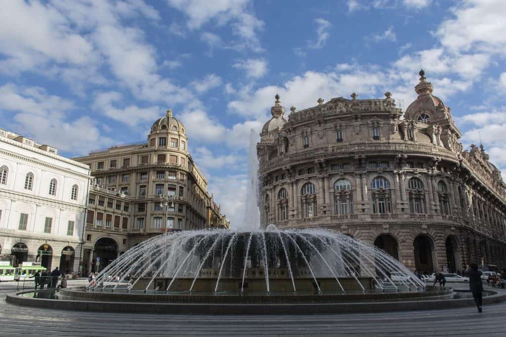 Beautiful historic city square with ornate architecture and a water fountain in the foreground, under a bright blue sky with scattered clouds, ideal for travel in Italy.