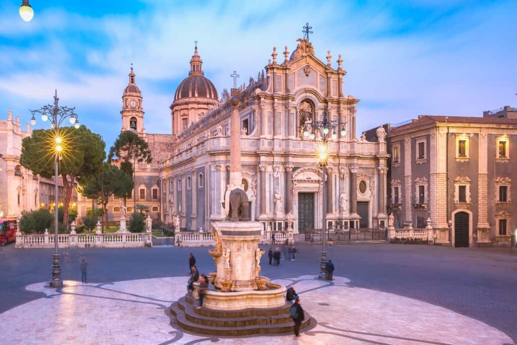 A cathedral with a baroque facade stands in a plaza with a central fountain, surrounded by historical buildings under a blue sky.