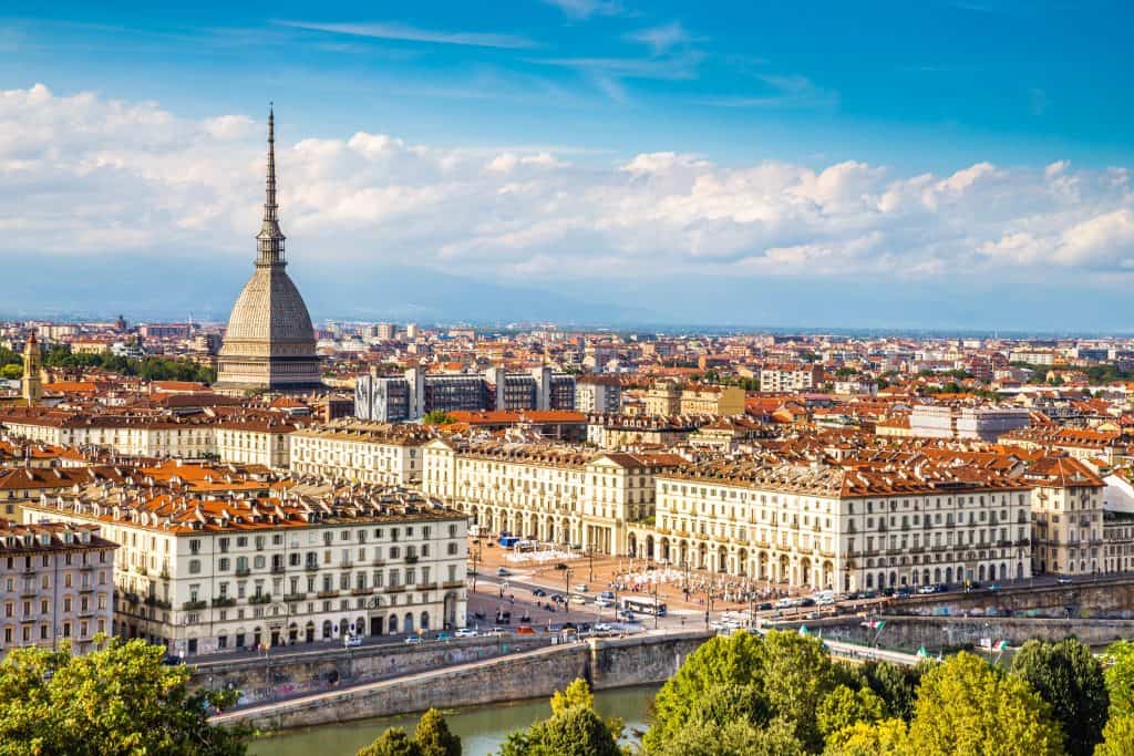 Vibrant aerial view of Turin, Italy, showcasing the Mole Antonelliana and historic cityscape with red-tiled roofs, scenic river, and lush greenery on a bright, sunny day.