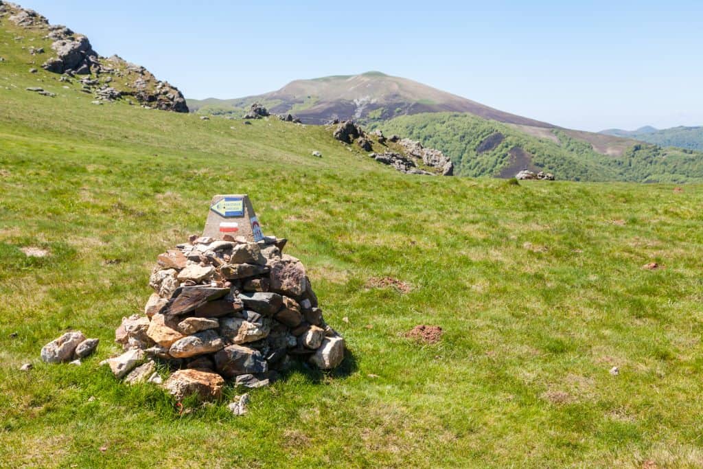 Cairn with trail marker on grassy hillside, mountain range in background under clear blue sky.